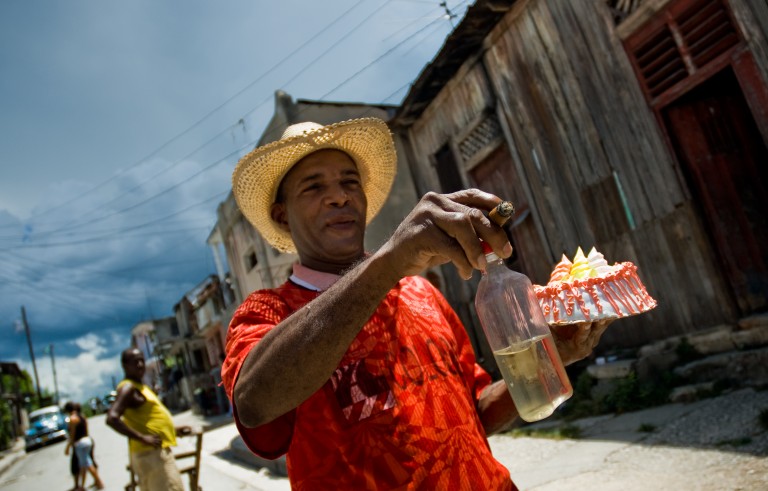 A Cuban man, holding a bottle of rum, smoking a cigar and carrying a cake, heads to a fiesta in Santiago de Cuba, Cuba. Photo by Jan Sochor via Getty Images