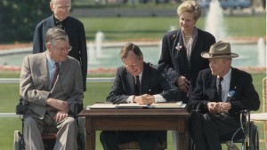 George Bush signs the Americans with Disabilities Act of 1990; standing left to right Reverend Harold Wilkie, Sandra Parrino of the National Council on Disability; seated left to right, Evan Kemp, Chairman of the Equal Employment and Opportunity Commission, George Bush, Justin Dart, Chairman of the 's Committee on the Employment of People with Disabilities. Washington DC, USA, 26 July 1990. (Photo by Fotosearch/Getty Images).