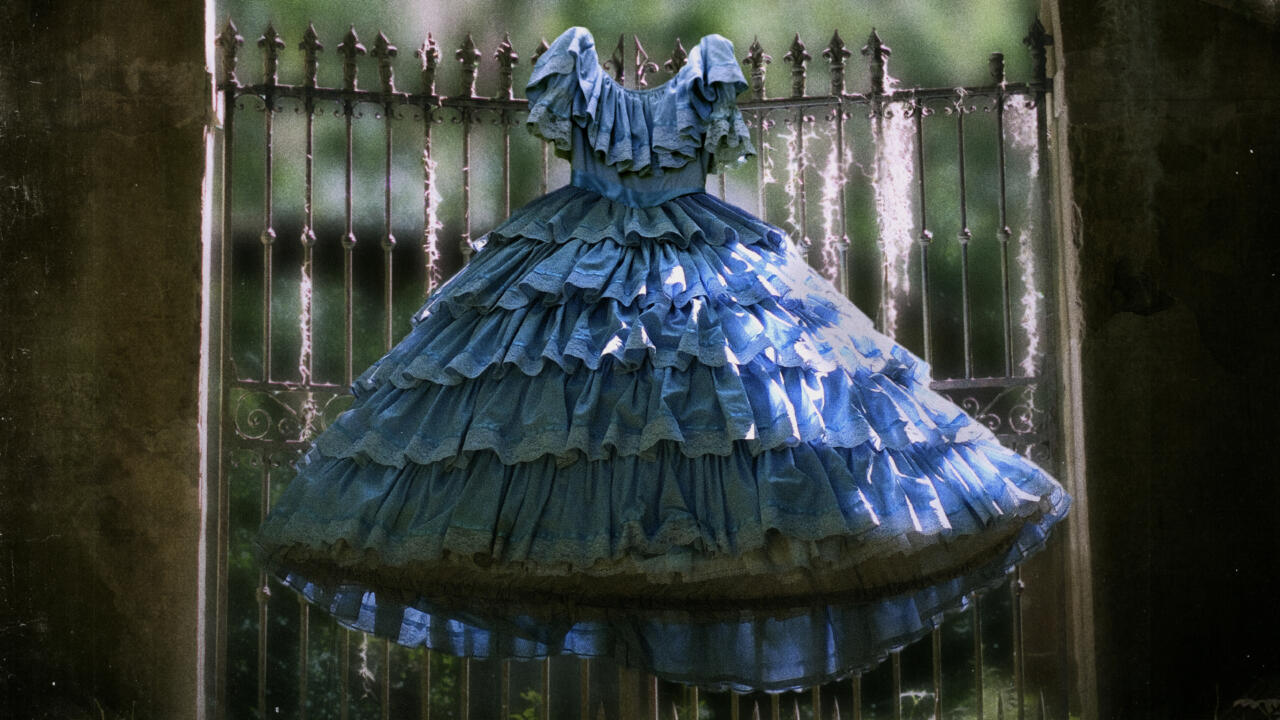 A headless blue antebellum ball gown stands before an aged wrought-iron gate in a Southern Gothic style