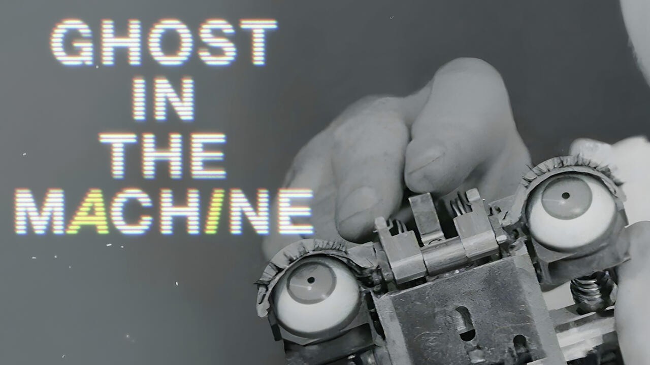 A person holds a small robot with round eyes. Overlaid, glitchy text reads “GHOST IN THE MACHINE.” The background is gray, and only part of the person’s face is visible.