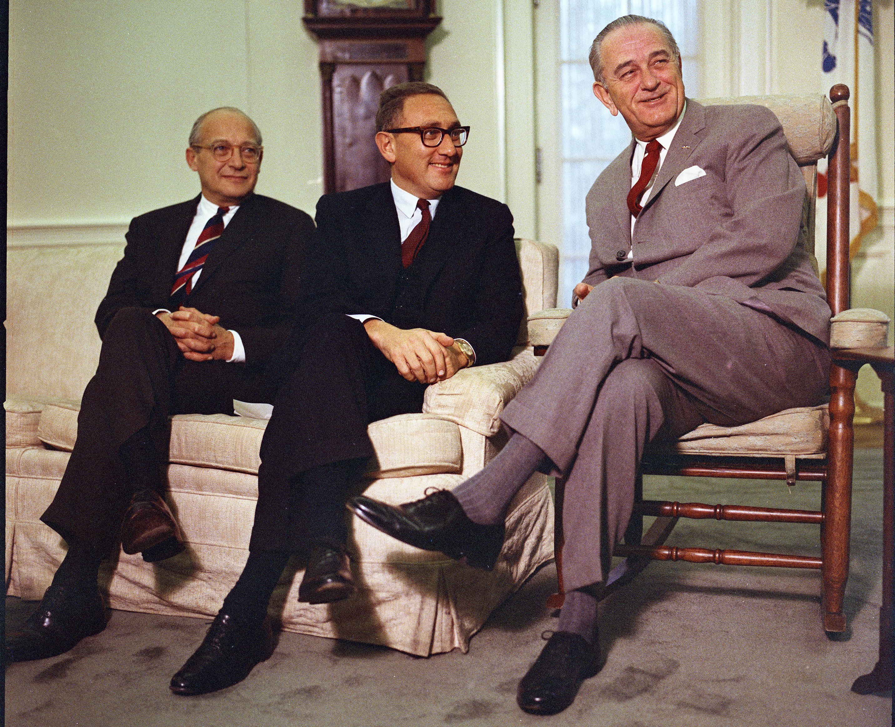 President Lyndon B. Johnson meets with Dr. Henry Kissinger and Walt Rostow. December 5, 1968. (National Archives) Three men sit together in a formal setting; two on a beige couch and one in a wooden chair. All are wearing suits and ties, smiling and appearing engaged in conversation. The atmosphere suggests a professional meeting or discussion.