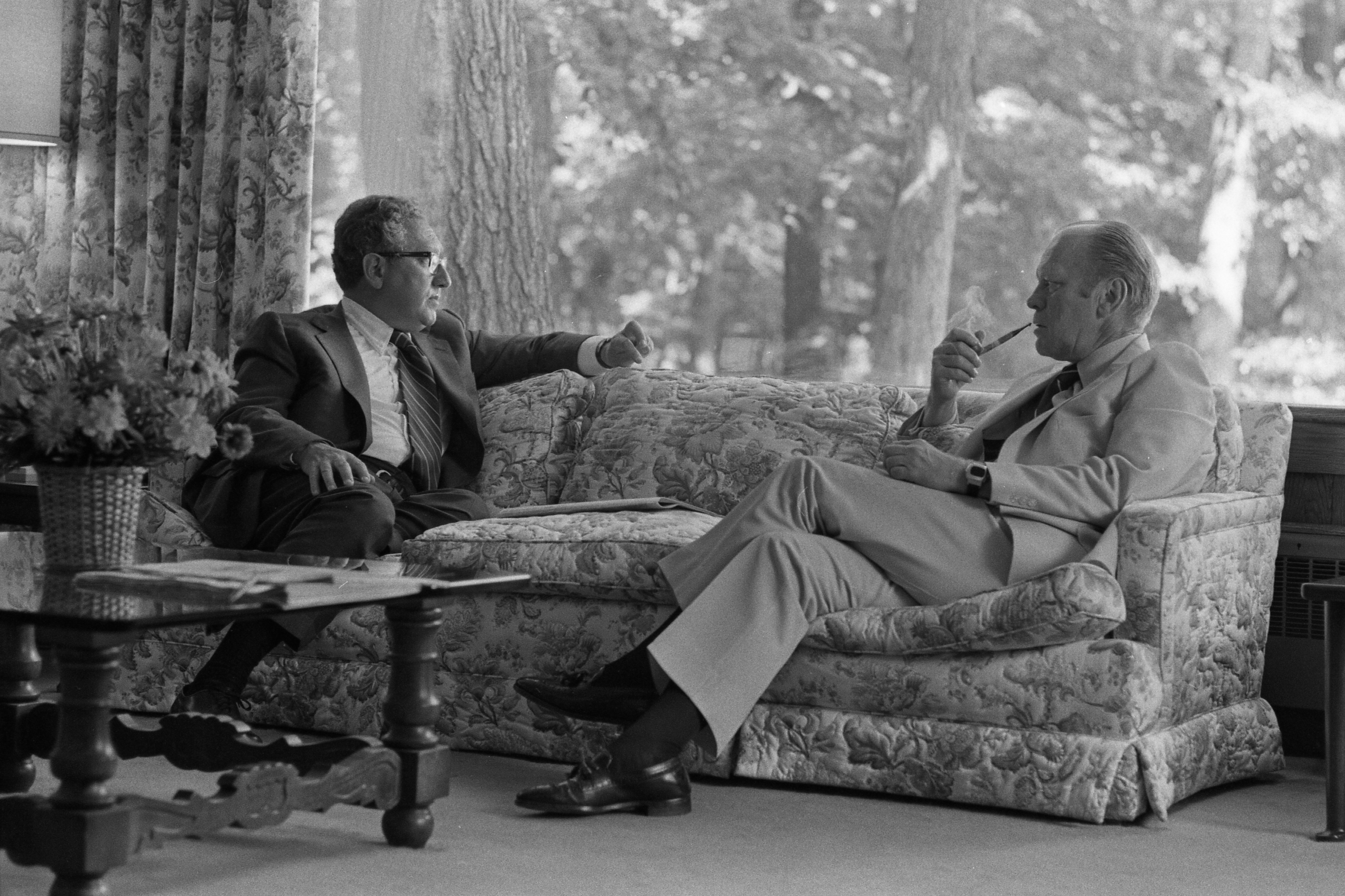 President Gerald Ford Meeting with Secretary of State and National Security Adviser Henry Kissinger at Camp David, July 5,1975. (National Archives) Two men sit on a patterned sofa in a relaxed, sunlit room, engaged in conversation. One gestures while speaking, and the other listens thoughtfully, holding a pipe. The setting appears informal, with large windows showing trees outside and a coffee table