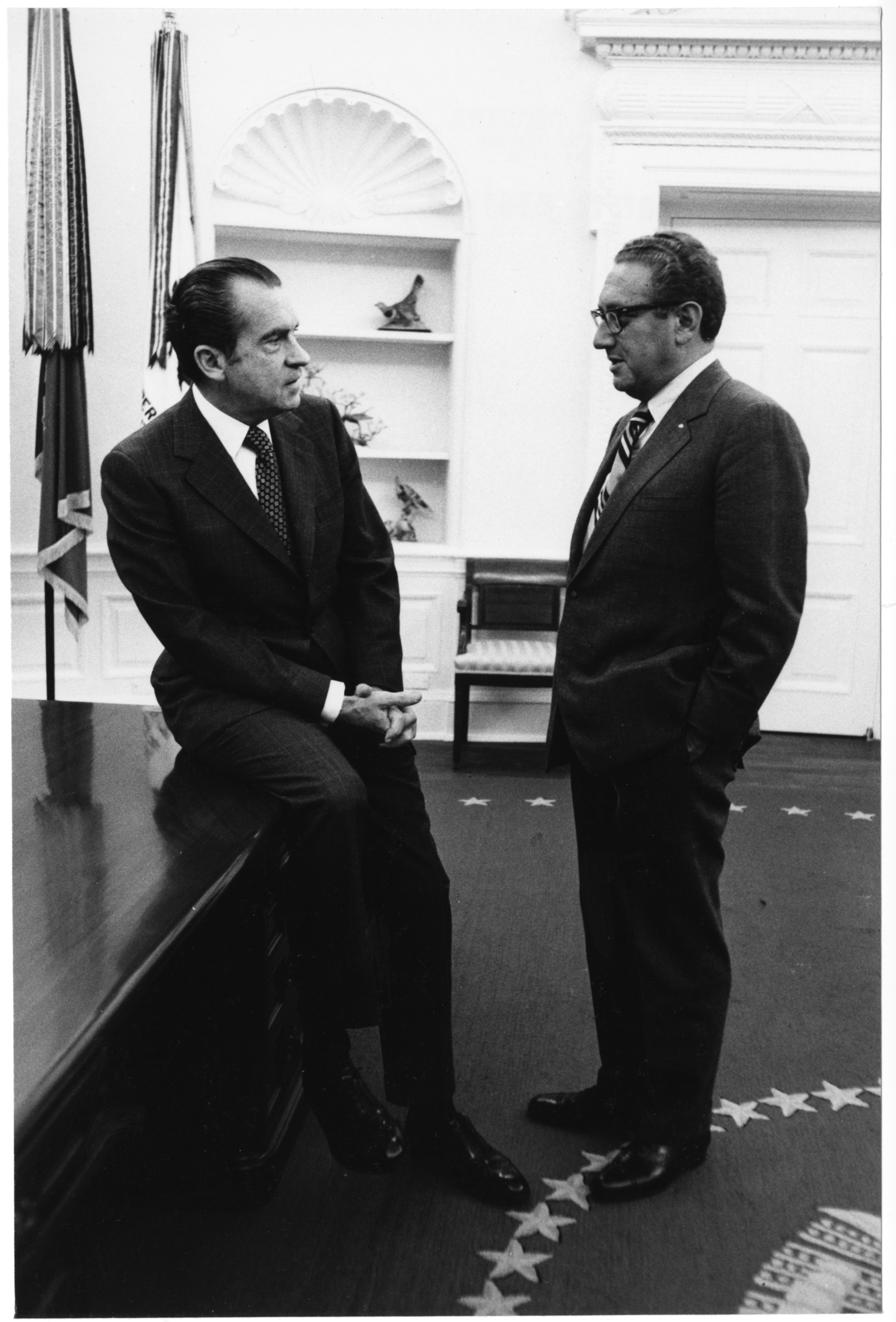 President Richard Nixon and National Security Advisor Dr. Henry Kissinger, 1972. (National Archives) Two men in suits have a serious conversation in an office. One sits on the edge of a large desk with his hands clasped, while the other stands with his hands in his pockets. The setting is formal, with flags, decorative shelves, and a carpet marked with s