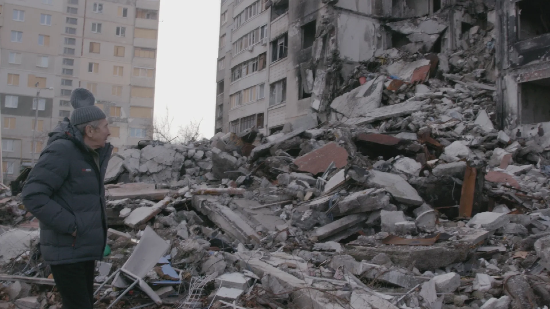 An old man dressed in winter clothes looks at the rubble of a destroyed building.