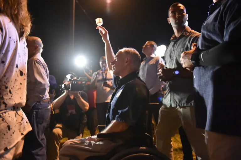 Texas Gov. Greg Abbott sits in a wheelchair and holds up a lit candle in a crowd at night.