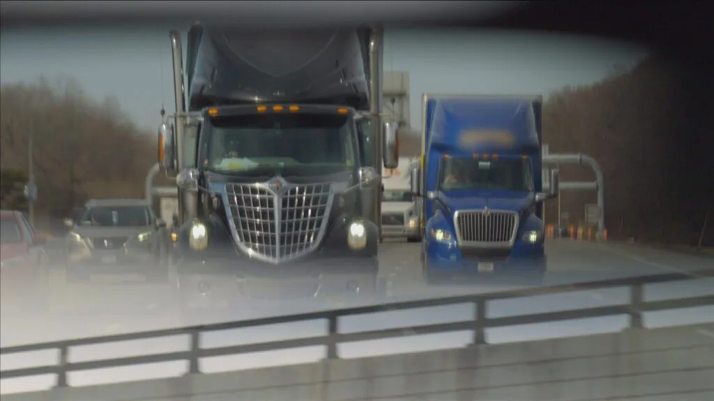 Trucks driving on a highway seen through a rearview mirror.