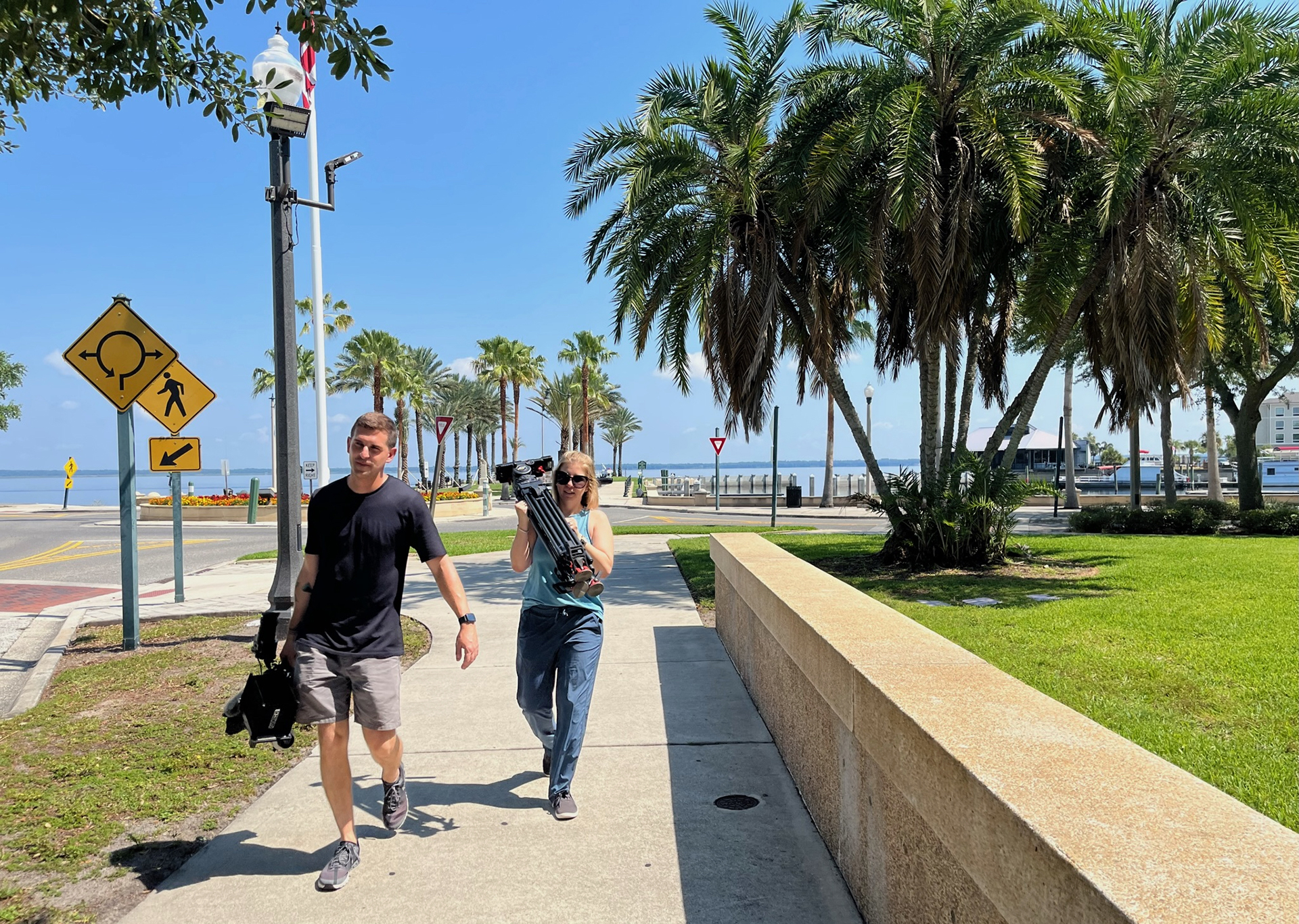 Isaac Mead-Long, left, and Cary Aspinwall carry camera equipment on a sunny day with palm trees and a body of water behind them.