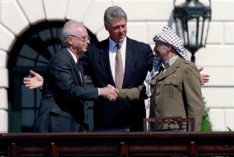 Former U.S. President Bill Clinton stands behind former Israeli Prime Minister Yitzhak Rabin and former PLO Chairman Yasser Arafat as they shake hands.