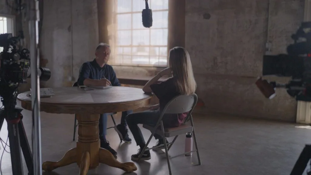 Two people sit at a table, and one person takes notes. There are two video cameras pointing at them.