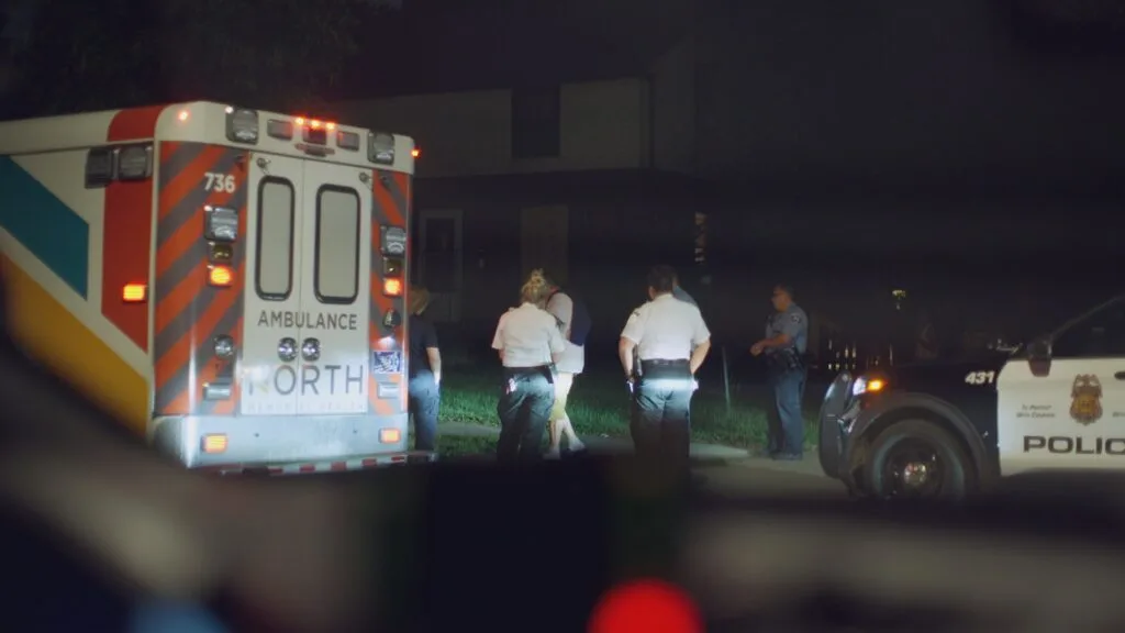 First responders and police stand near an ambulance and a police vehicle at night.