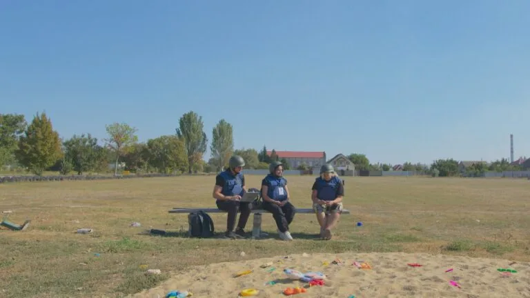 Three people wearing blue vests and helmets sit on a bench outdoors.