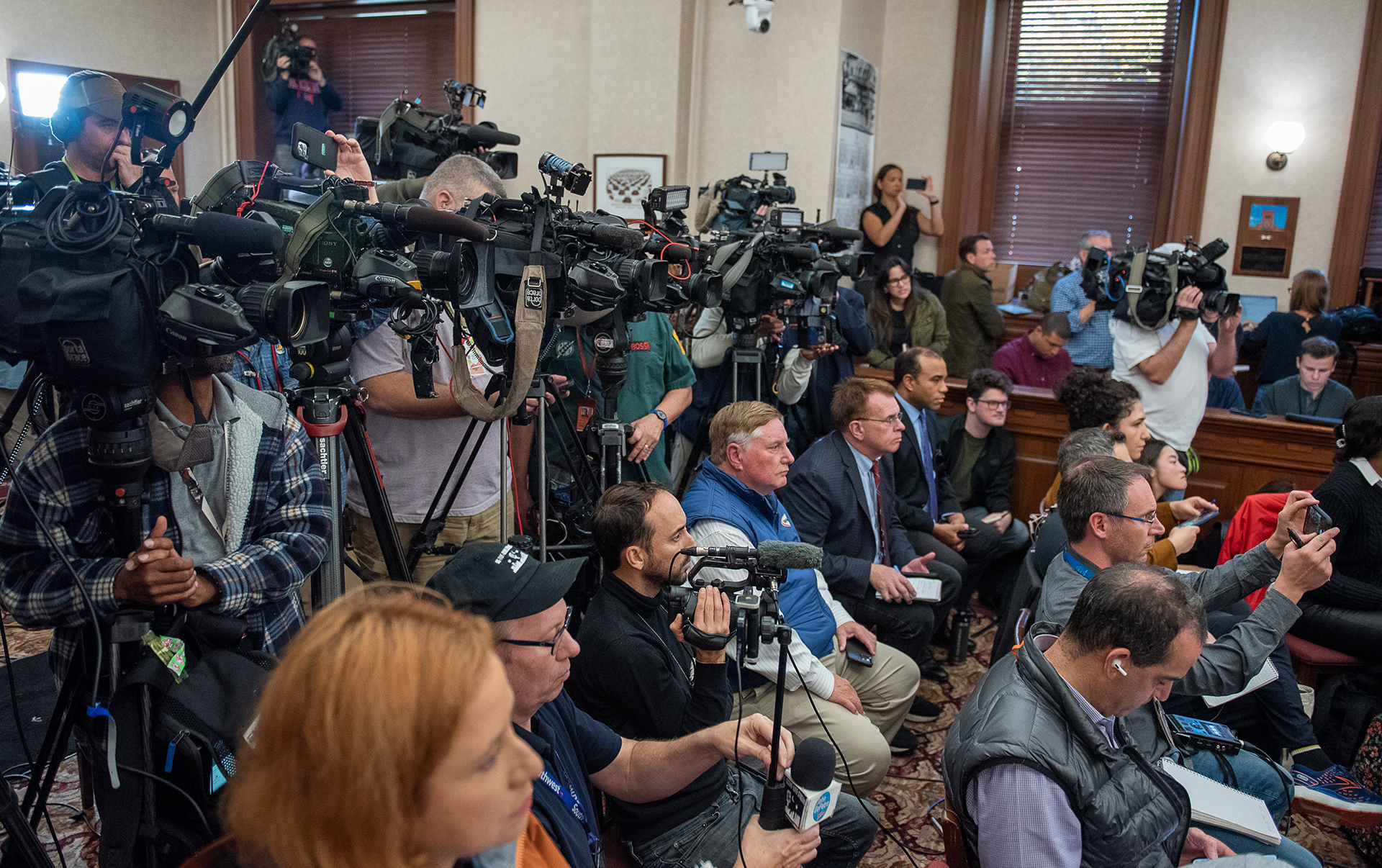 A crowd of reporters behind cameras on tripods or holding microphones, phones or notepads.
