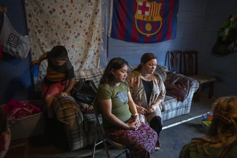Two women sit in a room with a young child behind them.