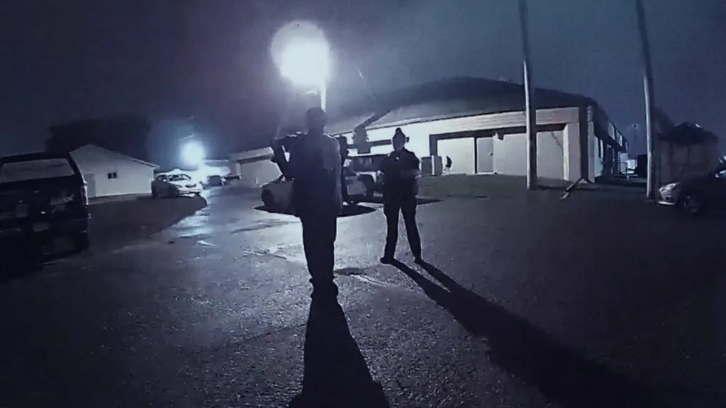 Two obscured figures shown at night under street lights.