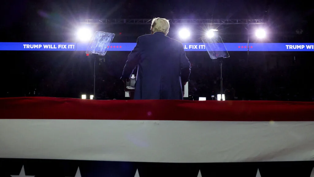 Donald Trump on the campaign trail speaking at a rally with his back to the camera and an electronic banner that says, "Trump will fix it."