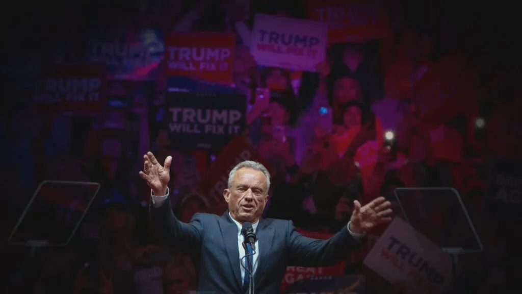 Robert F. Kennedy Jr. raising his hands at a campaign rally with people holding "Trump will fix it" signs in the background.