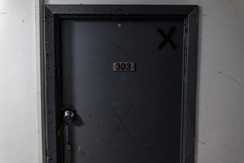 A door with three X’s drawn on it. The doorknob is broken and dangling.
