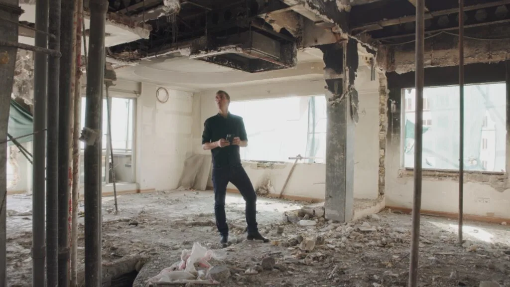 A man stands in the destroyed interior of a building holding a phone and looking up at the damaged ceiling.
