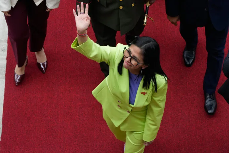 Delcy Rodriguez pictured from above wearing a lime green suit, standing on a red carpet and waving while three people walk behind her.