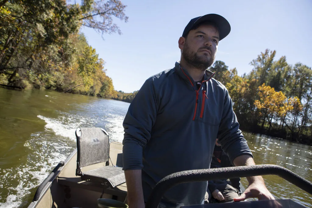 A man wearing a hat and blue fleece steers a boat on a river in broad daylight.