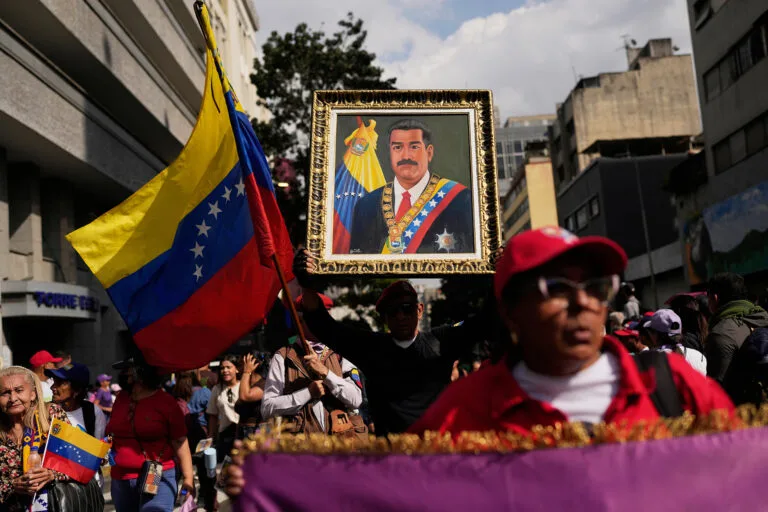 A scene of a crowd in a street, with a person holding up a painting of former Venezuelan leader Nicolas Maduro and and others holding up Venezuelan flags.