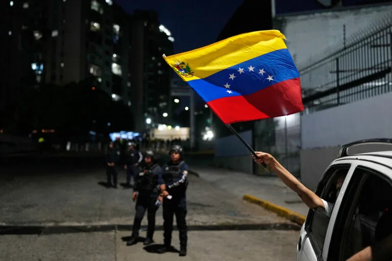 A person sticks their arm out of a passing vehicle waving a Venezuelan flag as law enforcement stand in the background.