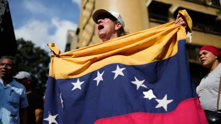 A person wearing a hat holds a Venezuelan flag and shouts.
