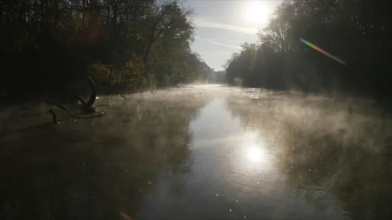 A river with mist rising off of it while the sun shines down.