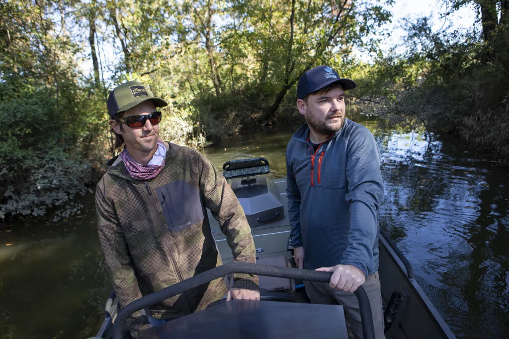 Two men wearing hats stand on a skiff in a river, with greenery on the river banks.