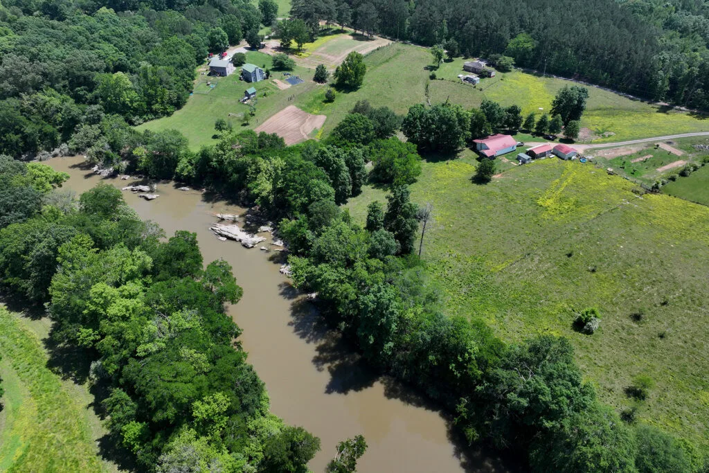 An overhead perspective of a muddy river with lush, green trees along the banks and houses nearby.
