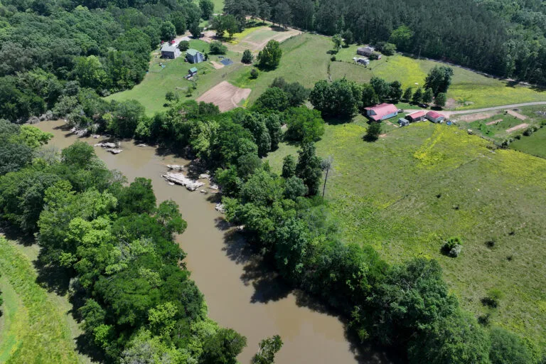 An overhead perspective of a muddy river with lush, green trees along the banks and houses nearby.