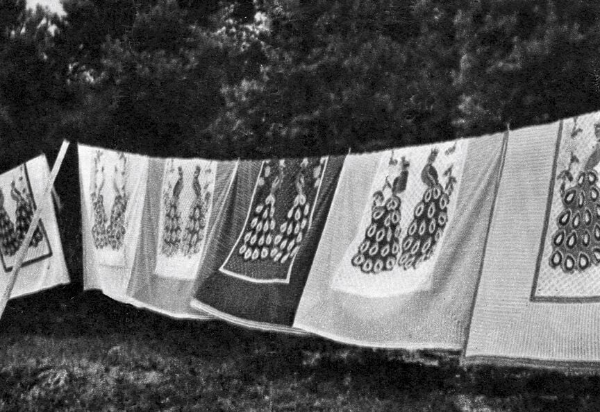 A black and white image of tufted quilts with peacock designs hanging on a clothesline outdoors.