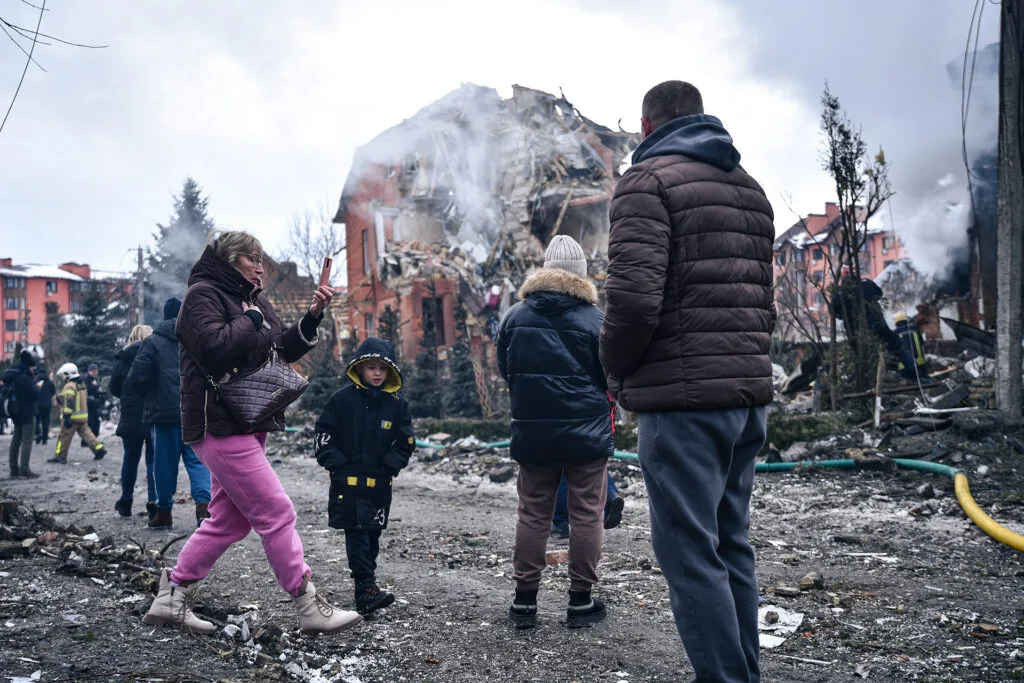 People and children stand on a street littered with debris, with first responders walking in the background. Behind the people is a destroyed building with half its structure crumbling and smoke emanating from it.