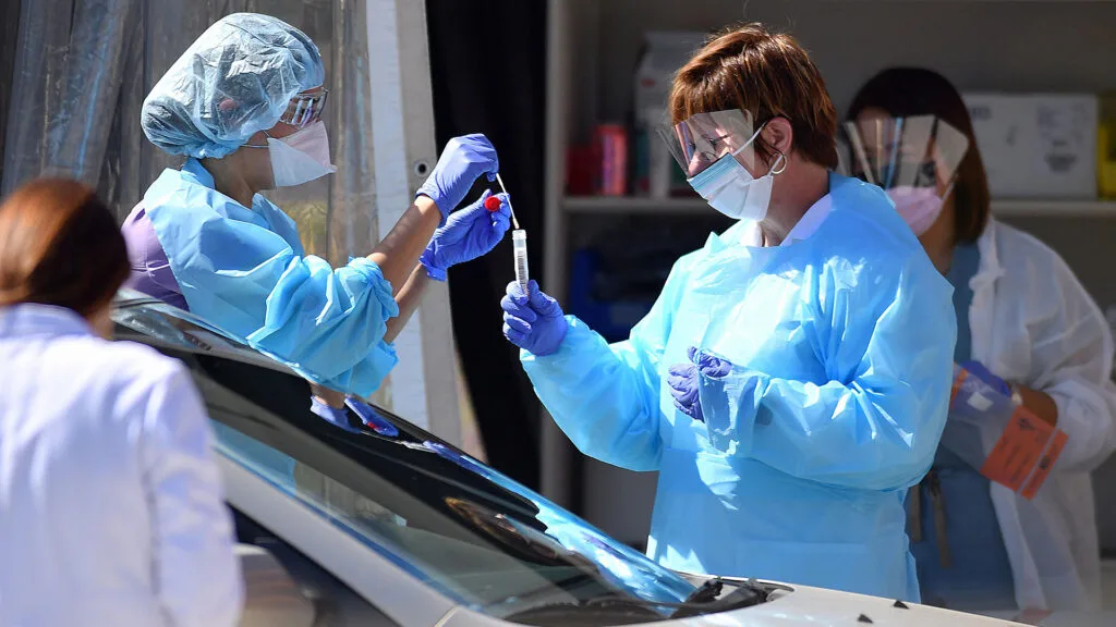 Medical workers wearing masks, gloves and protective gear stand next to a car holding a swab and a tube.