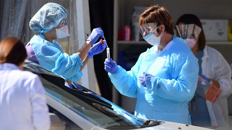 Medical workers wearing masks, gloves and protective gear stand next to a car holding a swab and a tube.