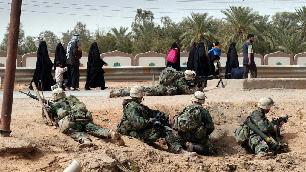 Men, women and children walk on a road in the background, while armed soldiers wearing helmets and backpacks crouch in a ditch on the side of the road.