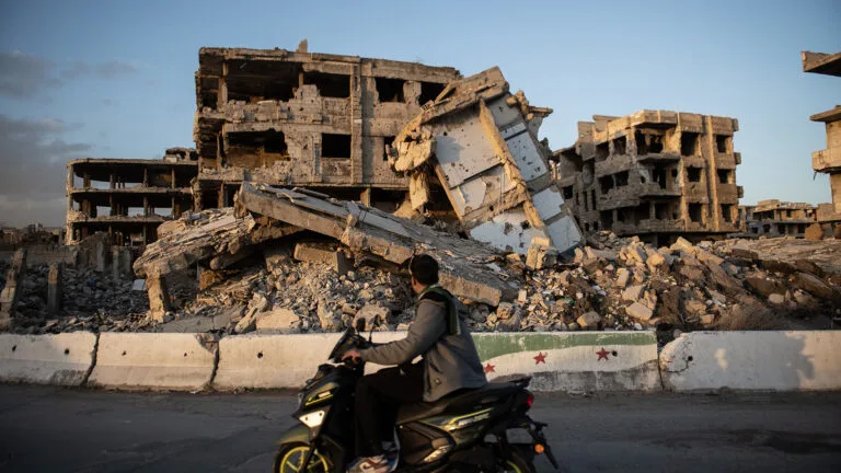 A destroyed building in the background as a man rides by on a scooter.