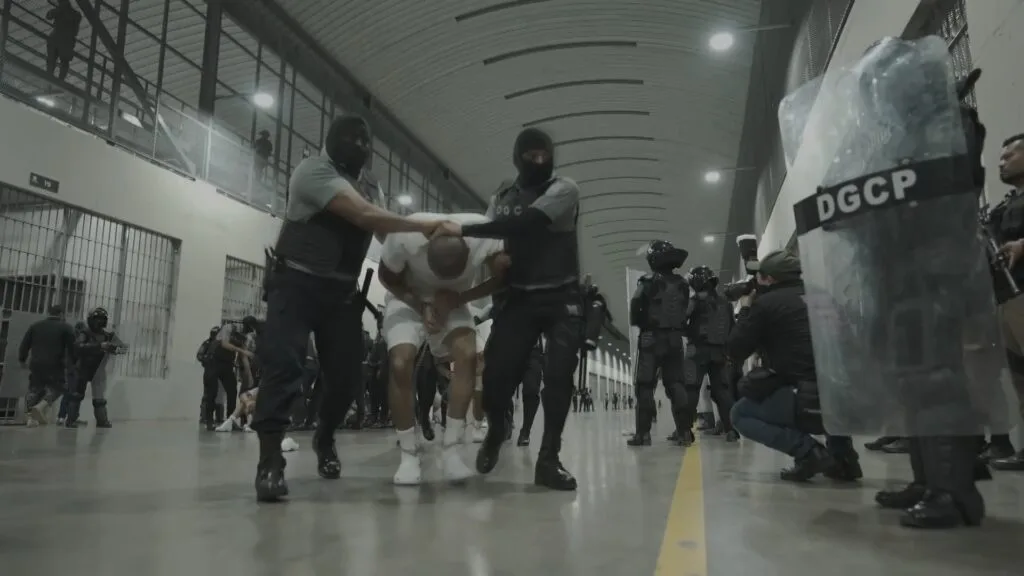 Several armored guards stand inside the Salvadoran prison CECOT and two guards wearing balaclavas restrain a prisoner as they walk him elsewhere.