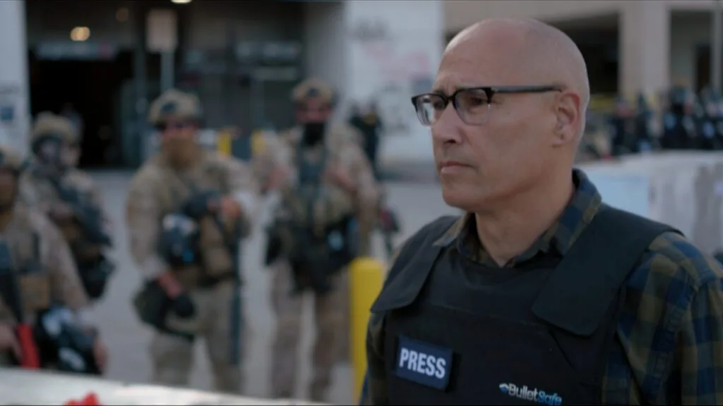 Correspondent A.C. Thompson stands in the foreground wearing a bulletproof vest that says "press" on it, while federal agents stand in the background.
