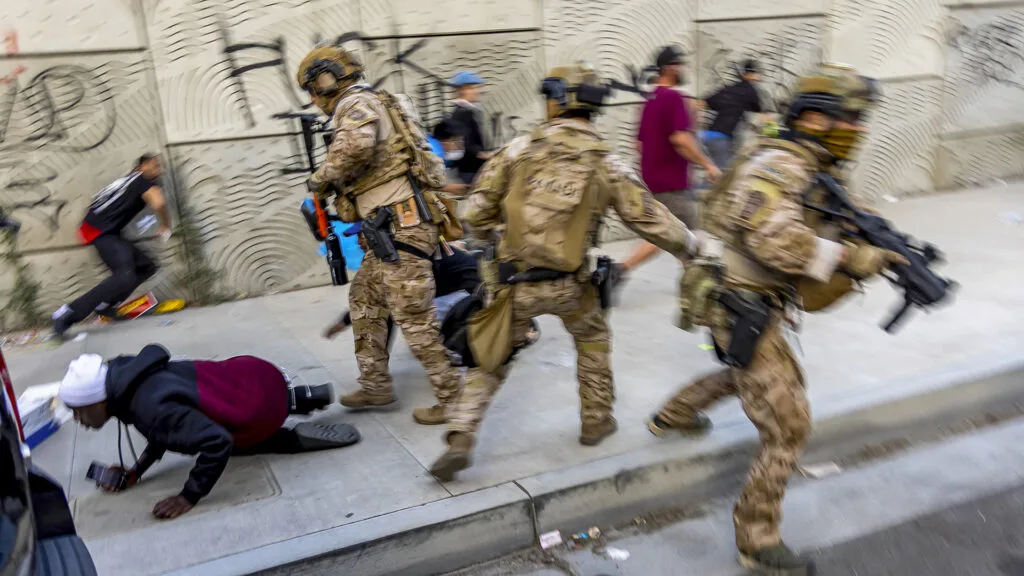 In a hectic scene, three officers with guns, helmets and camouflage gear are a blur of motion on a sidewalk, and all three are moving in different directions. Behind and beside them are people in street clothes, including one person who has fallen on the sidewalk. They are holding a phone and an agent is standing over them.