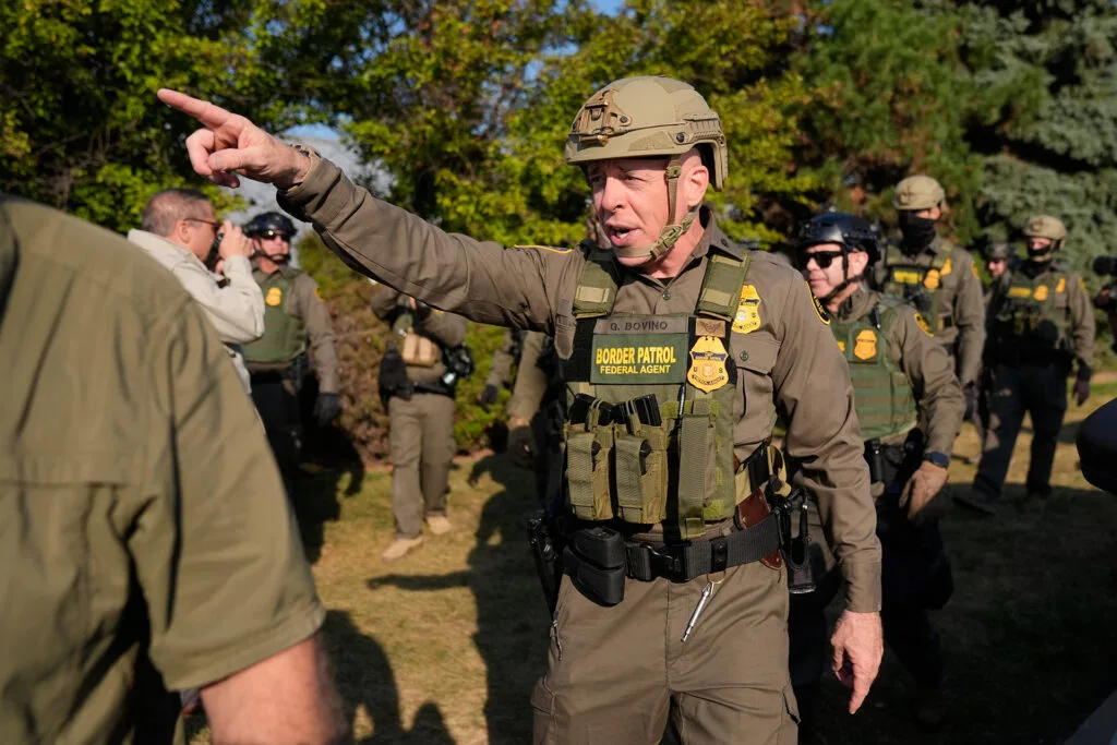 A man wearing a brown Border Patrol uniform, helmet and vest points off screen.