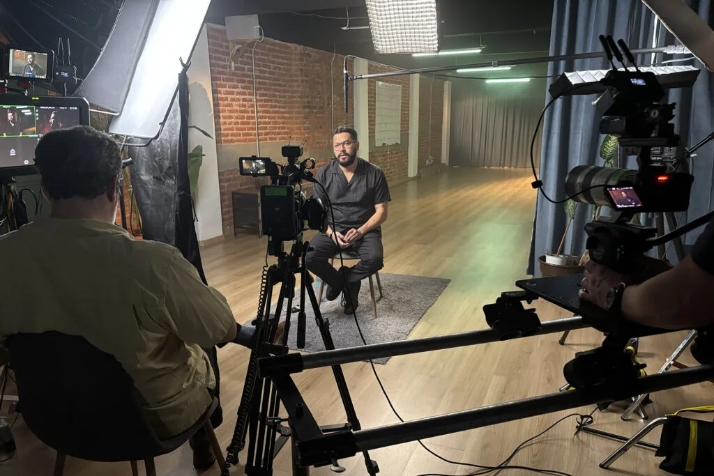 Óscar Martínez being interviewed by a man viewed from the back in a room with several cameras and monitors in the foreground.