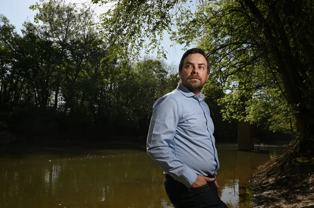 A man in a button-down shirt stands on the banks of a river.