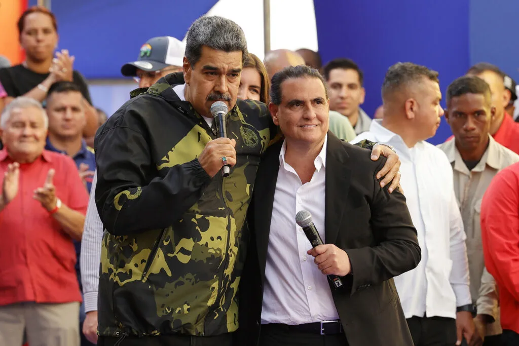 Venezuelan leader Nicolás Maduro stands wearing a windbreaker and holding a mic, with his arm around a man wearing a suit and also holding a mic.