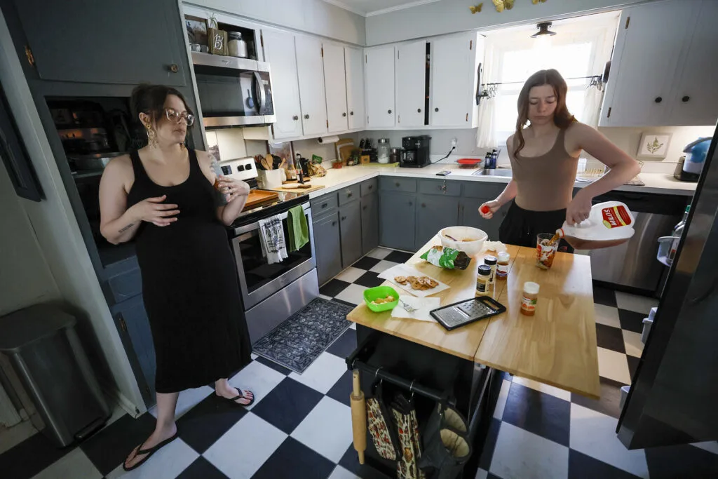 A woman and a teenage girl stand in a kitchen, the girl pouring a drink from a gallon container into a tall glass.