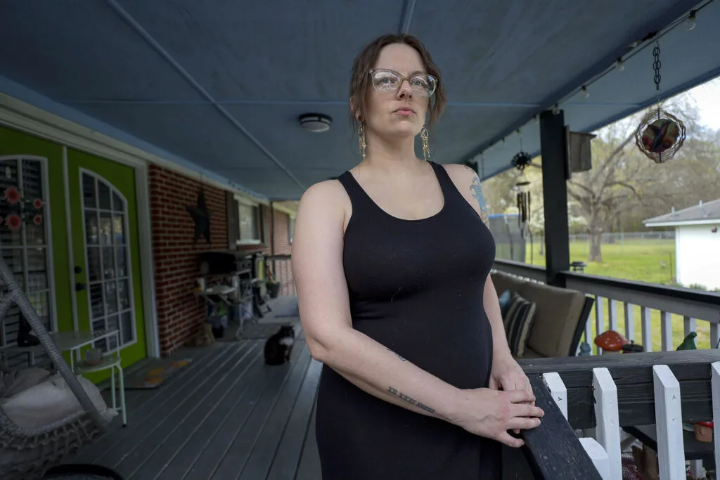 A woman wearing a sleeveless dress, dangling earrings and glasses stands on a porch.