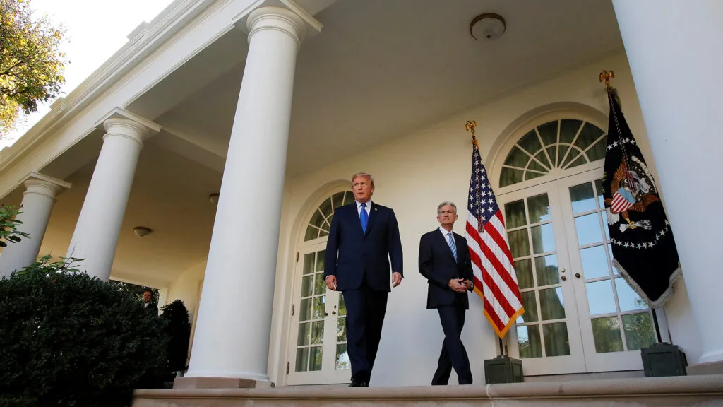 President Donald Trump and Federal Reserve Chair Jerome Powell seen walking in front of a White House exterior with flags hanging behind them.