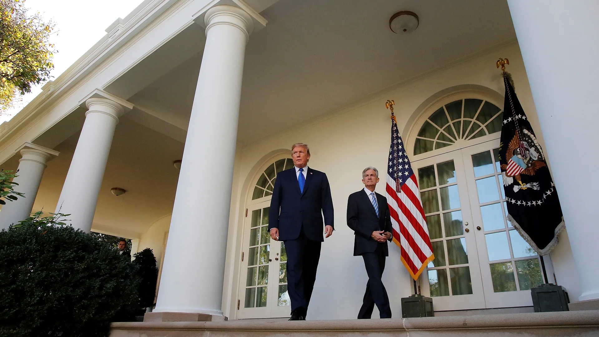 President Donald Trump and Federal Reserve Chair Jerome Powell seen walking in front of a White House exterior with flags hanging behind them.