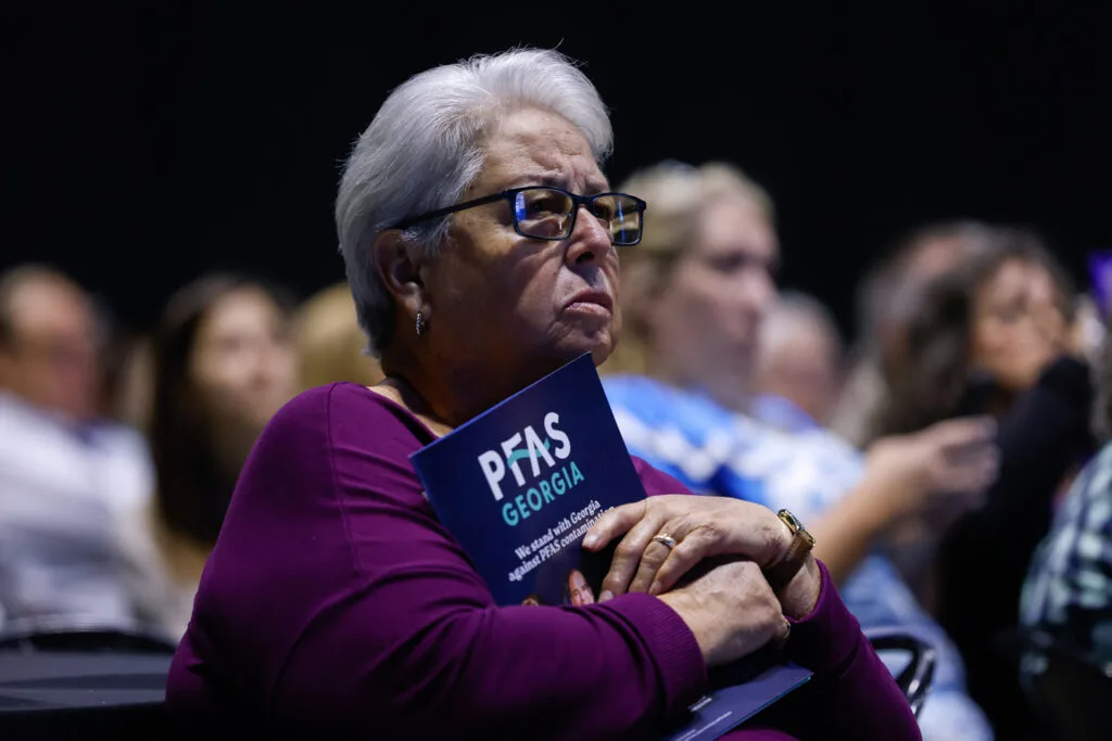 A silver-haired woman wearing glasses looks serious while holding a pamphlet that says "PFAS Georgia."