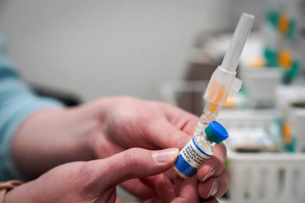 The hands of a nurse holding a vial of a dose of the measles, mumps and rubella vaccine and an unopened syringe.